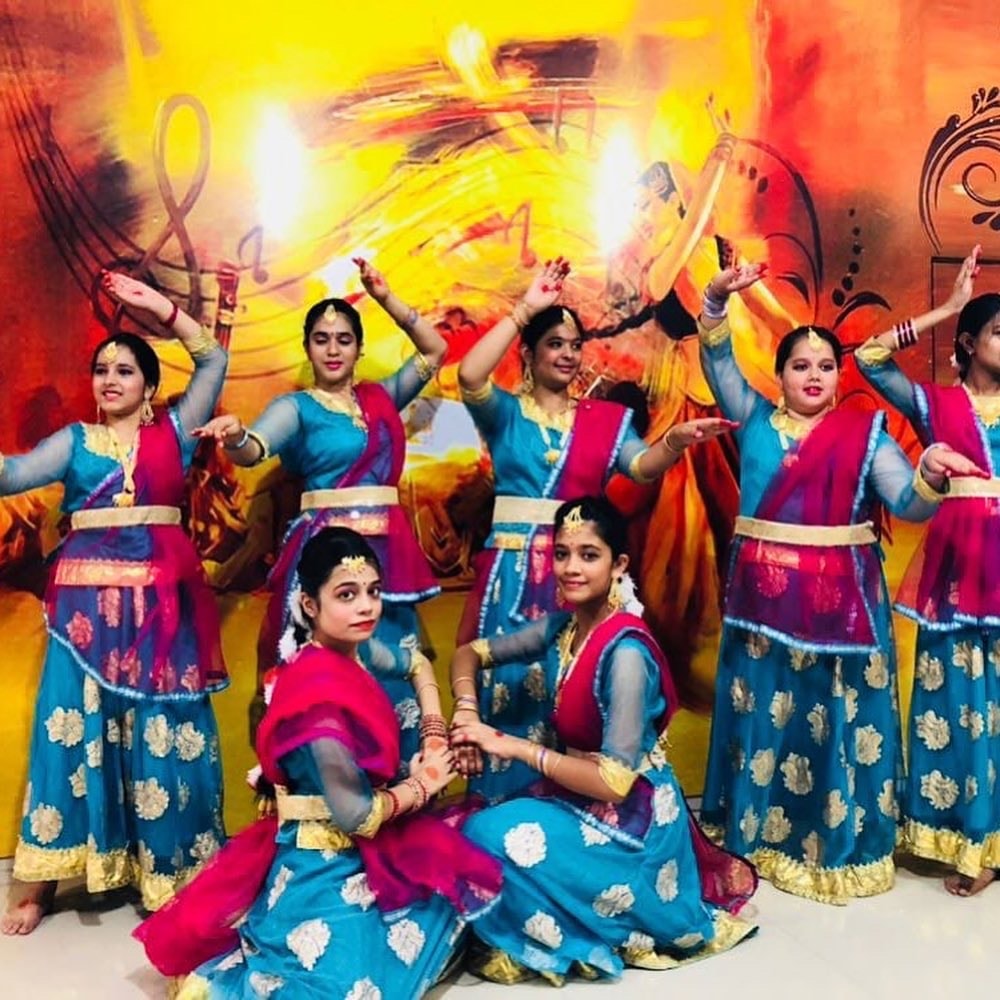 Group of eight dancers in teal and pink costumes performing against a vibrant painted backdrop